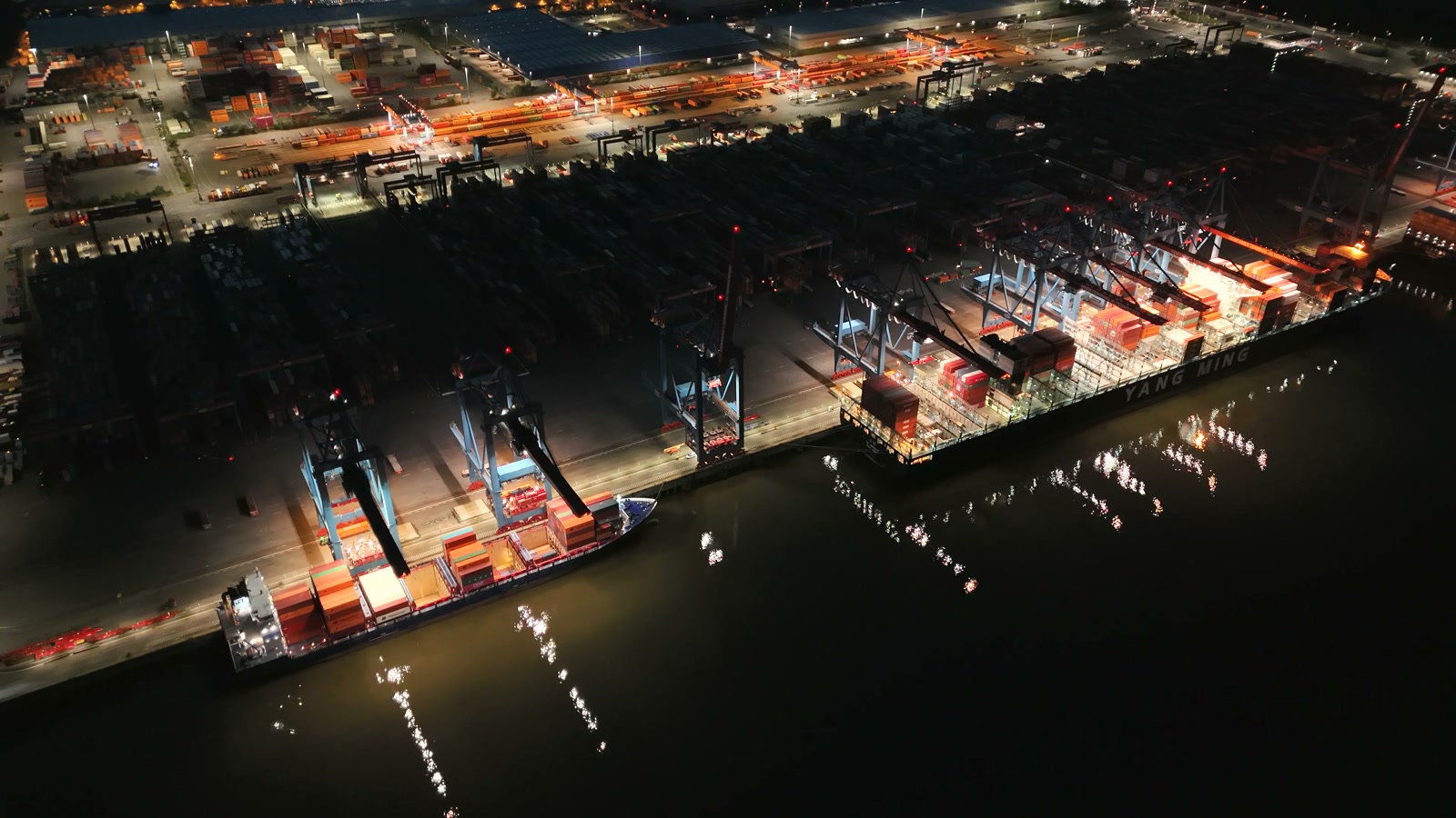 Night aerial of a container ship at berth under active gantry cranes.