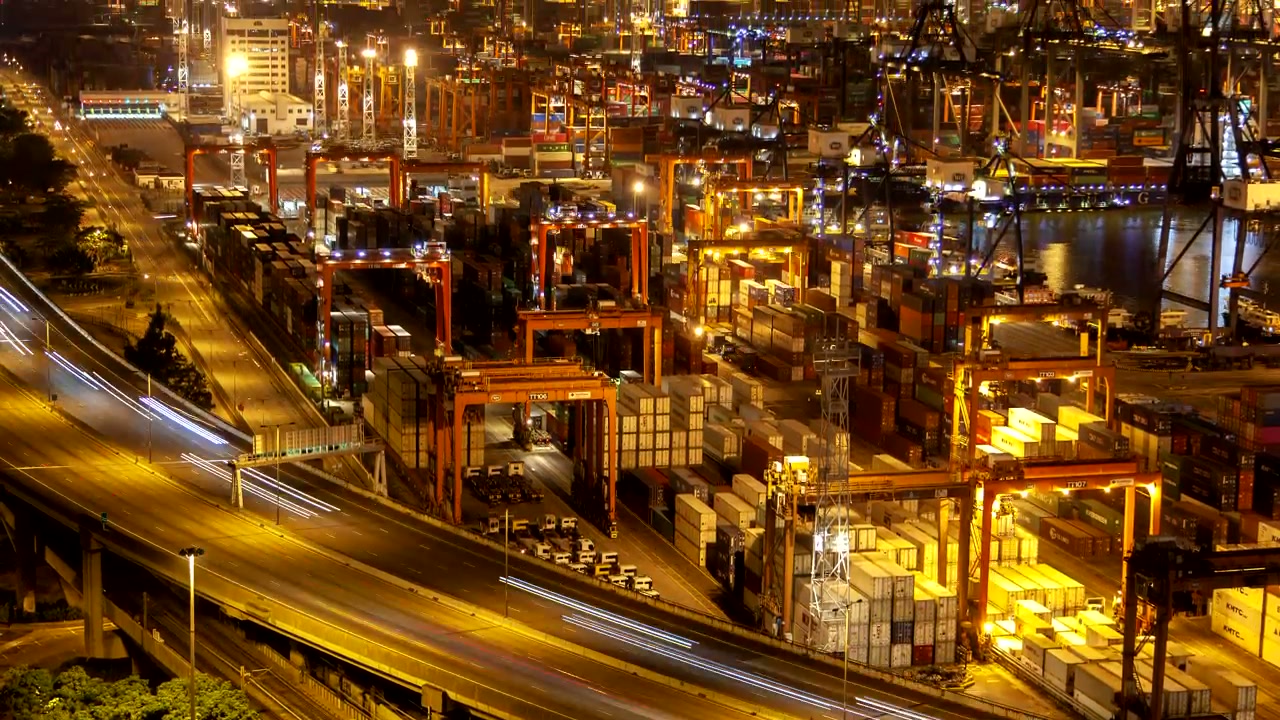Night aerial of a container terminal with gantry cranes beside a lit highway.