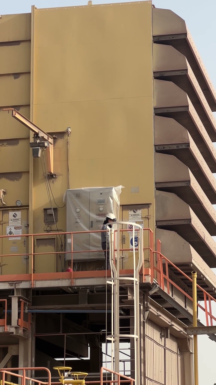 Technician in hard hat on a plant platform inspecting outdoor equipment.